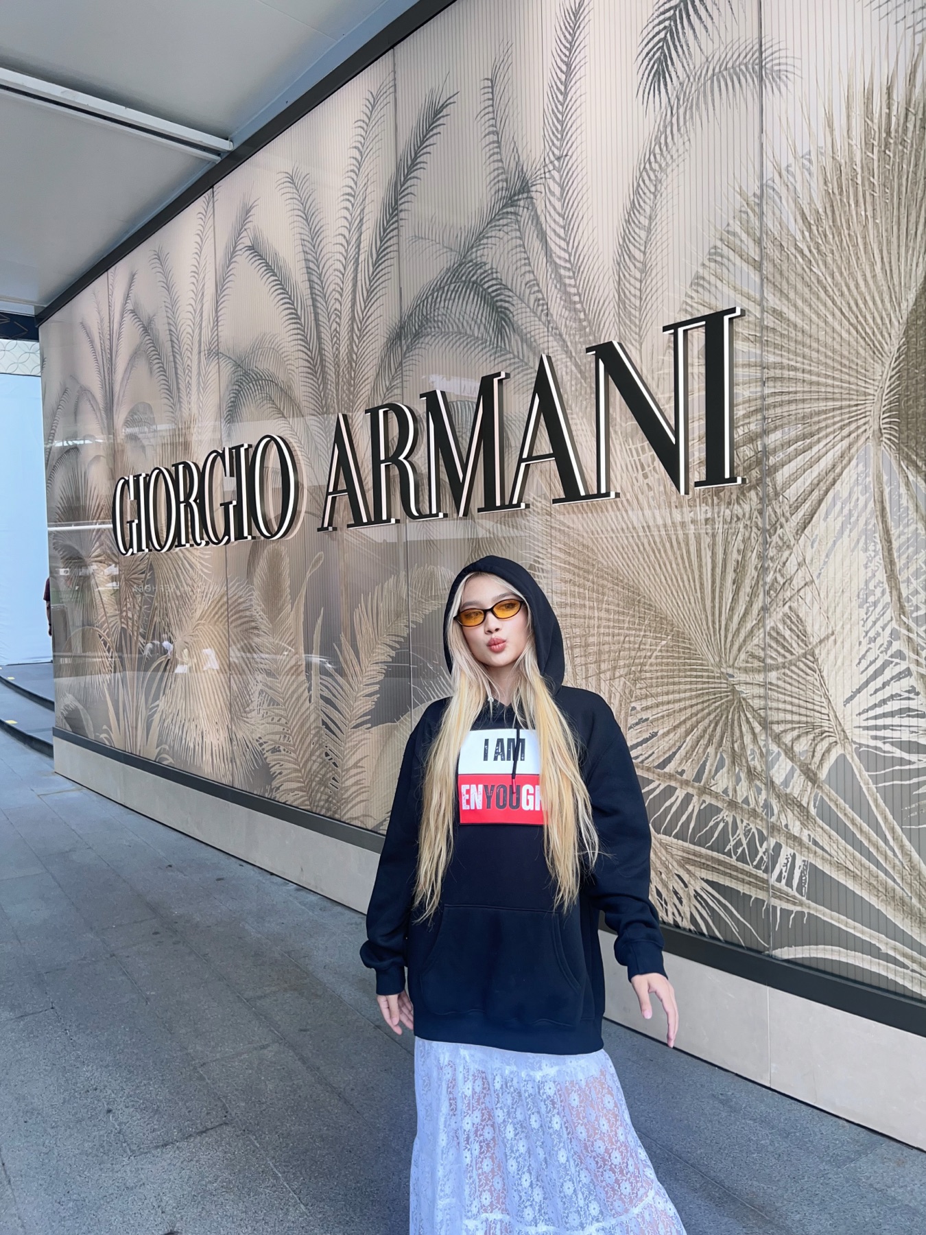 Model in the I Am Enyough hoodie, tinted sunglasses, standing in front of the Giorgio Armani storefront